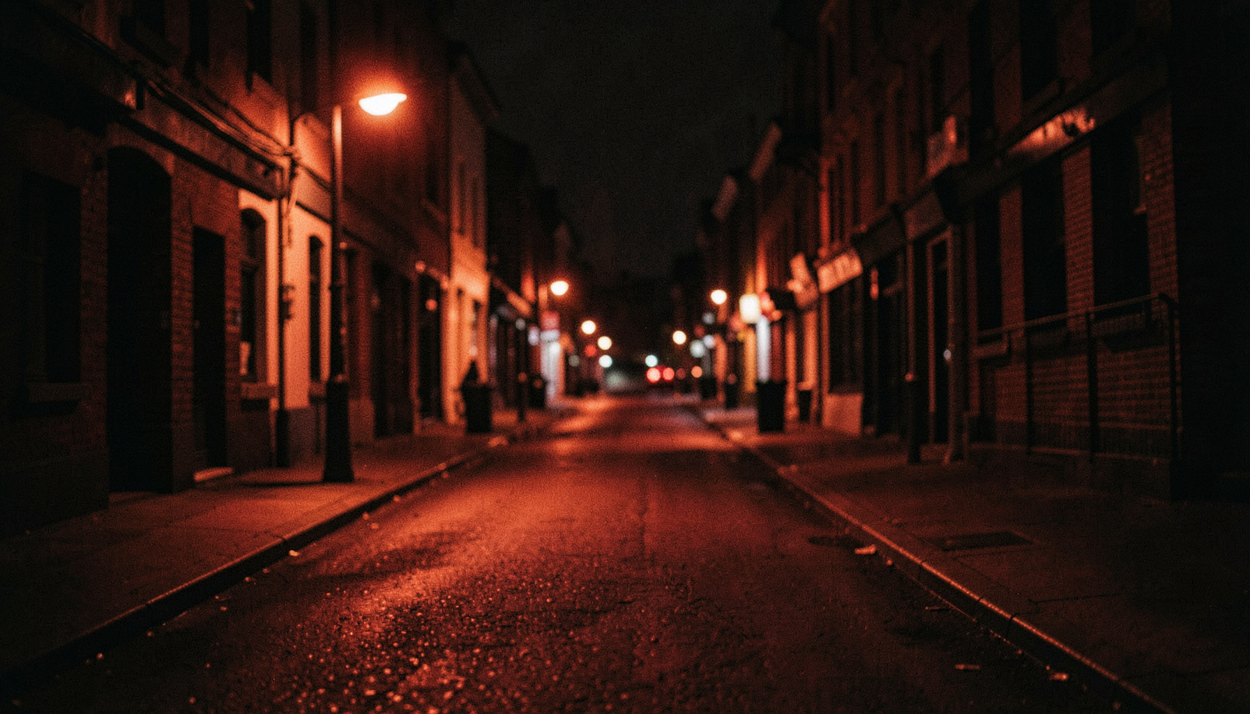 Soft cinematic frame of a quiet urban street at night, deep shadows and warm sodium lighting